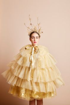 a woman in a yellow dress with a gold crown on her head, standing against a pink background