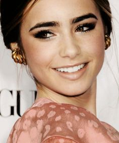 a close up of a person wearing gold earrings and a pink dress with black eyeliners