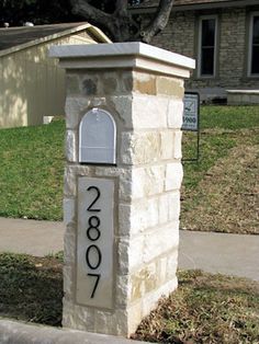 a stone mailbox on the sidewalk in front of a house with grass and trees