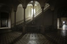 an empty hall with stairs and tiled floors in the dark, lit by sunlight coming through two windows