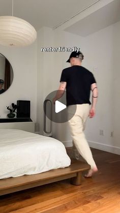 a man walking across a wooden floor next to a bed in a room with white walls