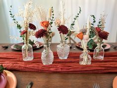 four clear vases with flowers are sitting on a red table cloth near plates and utensils