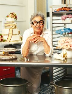 an older woman sitting at a table in front of some cakes and other desserts