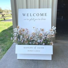 a welcome sign sitting on top of a cement slab in front of a building with flowers growing out of it