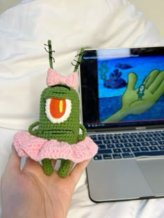 a hand holding a small crocheted toy next to a laptop