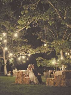 a couple sitting on hay bales under a tree at night with lights strung from the branches