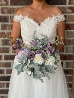 a woman wearing a wedding dress holding a bouquet of flowers and greenery in front of a brick wall