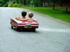 two young boys riding in the back of an old red convertible car on a paved road