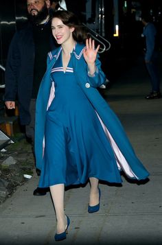 a woman in a blue dress is walking down the street with her hand up and smiling