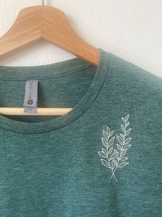 a green t - shirt with white leaves on the front and back, hanging on a wooden hanger