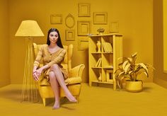 a woman sitting in a yellow chair next to a lamp and bookshelf with pictures on the wall