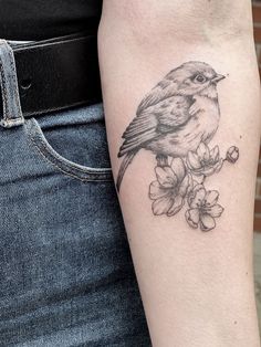 a bird sitting on top of a branch with flowers in it's beak, next to a woman's arm