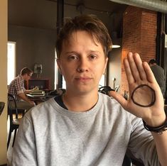 a man sitting at a table holding his hand up to the camera with another person in the background