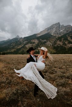 a bride carrying her groom in the middle of a field with mountains in the background