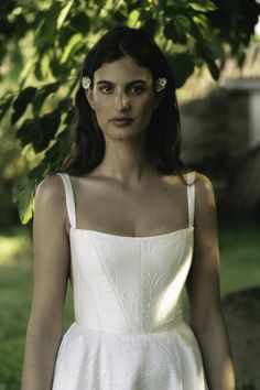 a woman wearing a white dress standing in front of a tree with her hands on her hips