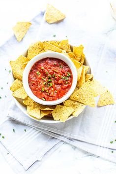 a white bowl filled with salsa surrounded by tortilla chips on top of a napkin