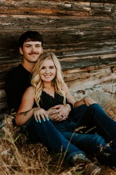 a man and woman sitting next to each other in front of a log wall with grass