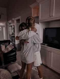 two women hugging each other in the middle of a living room with white walls and furniture