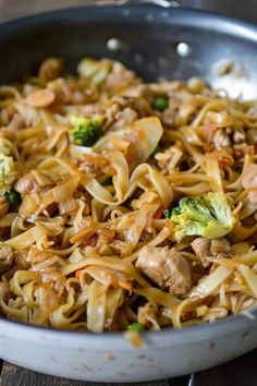 a skillet filled with noodles, meat and broccoli on top of a wooden table