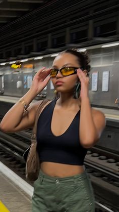 a woman wearing sunglasses standing in front of a train platform with her hand on her head