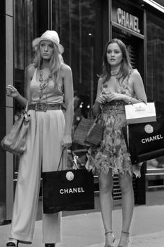 two women are standing on the sidewalk with shopping bags in hand and one is holding a chanel bag