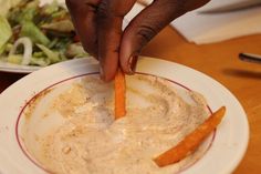 a person is dipping some food into a bowl with carrots on the side and salad in the background