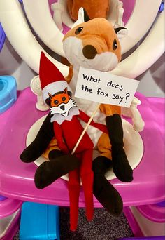 a stuffed animal sitting on top of a toilet seat holding a sign that says, what does the fox say?