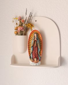 a statue of the virgin mary on a white shelf with flowers in a vase next to it