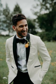 a man in a tuxedo smiles as he stands in a field with wildflowers