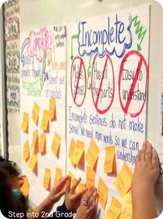 two children are writing on a bulletin board with sticky notes attached to the wall and another child is pointing at it