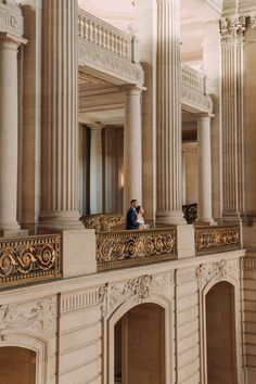 two people standing on the balcony of a large building with columns and gold trimmings