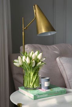 a table with a lamp, book and flowers on it in front of a couch