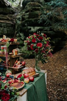 a table topped with fruit and flowers next to a forest filled with lots of trees