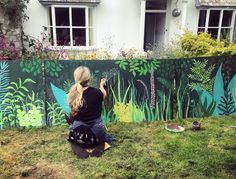 a woman is painting a mural on the side of a house with grass and flowers