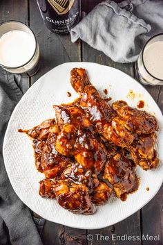 a white plate topped with chicken wings covered in bbq sauce next to two glasses of beer