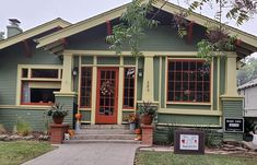 a small green house with red trim and two large windows on the second floor, has potted plants in front of it