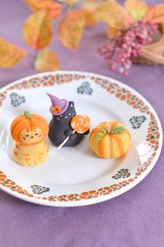 three small pumpkins are on a plate with two cats and one cat is holding a candy lollipop