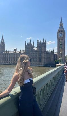 a woman standing on the side of a bridge next to a body of water with a clock tower in the background