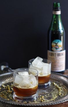 two glasses filled with ice sitting on top of a metal tray next to a bottle