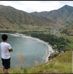 a man standing on top of a lush green hillside next to a lake and mountains