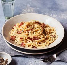 a white bowl filled with pasta and bacon on top of a table next to a glass of water