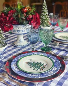 the table is set for christmas dinner with blue and white plates, silverware, red flowers and greenery