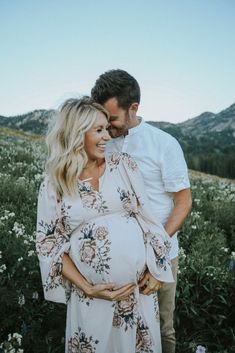 a pregnant couple standing in a field with mountains in the background and flowers all around them