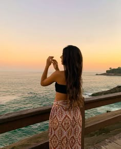 a woman standing on top of a wooden pier next to the ocean at sunset with her hand in her mouth