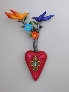 a red heart shaped vase with flowers and birds on it's top, hanging from a branch