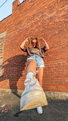 a woman leaning against a brick wall with her foot on the ground and wearing white sneakers