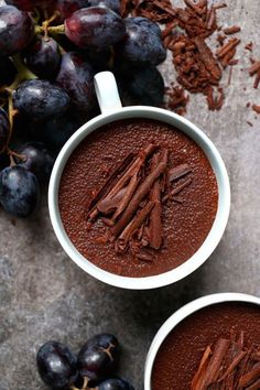 two mugs filled with chocolate next to some grapes and cinnamon on a table top