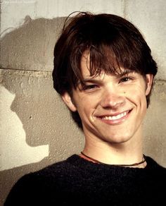 a young man smiling in front of a cement wall with his hair pulled back and wearing a black sweater