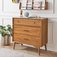 a wooden chest of drawers in a white room with plants and books on the shelves