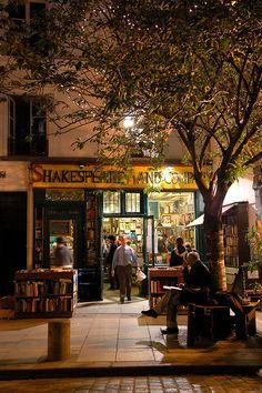 people are sitting on benches in front of a book store at night with the lights on